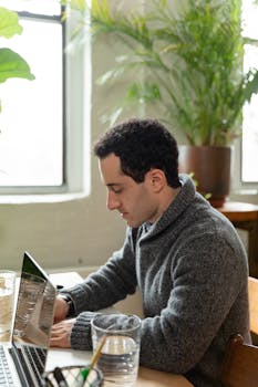 Focused man working on a laptop surrounded by plants in a bright office setting.