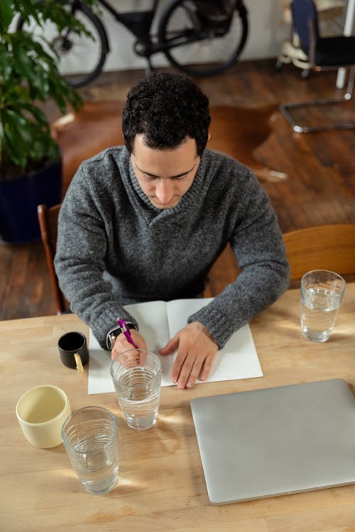 Man Sitting at the Table with a Laptop and Writing on Paper 