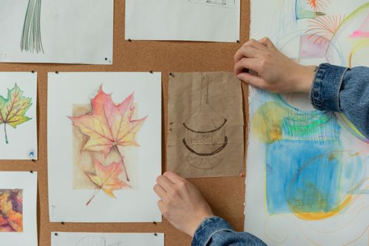 Close-up of hands pinning colorful leaf drawings on a corkboard, creating a cozy artistic atmosphere.
