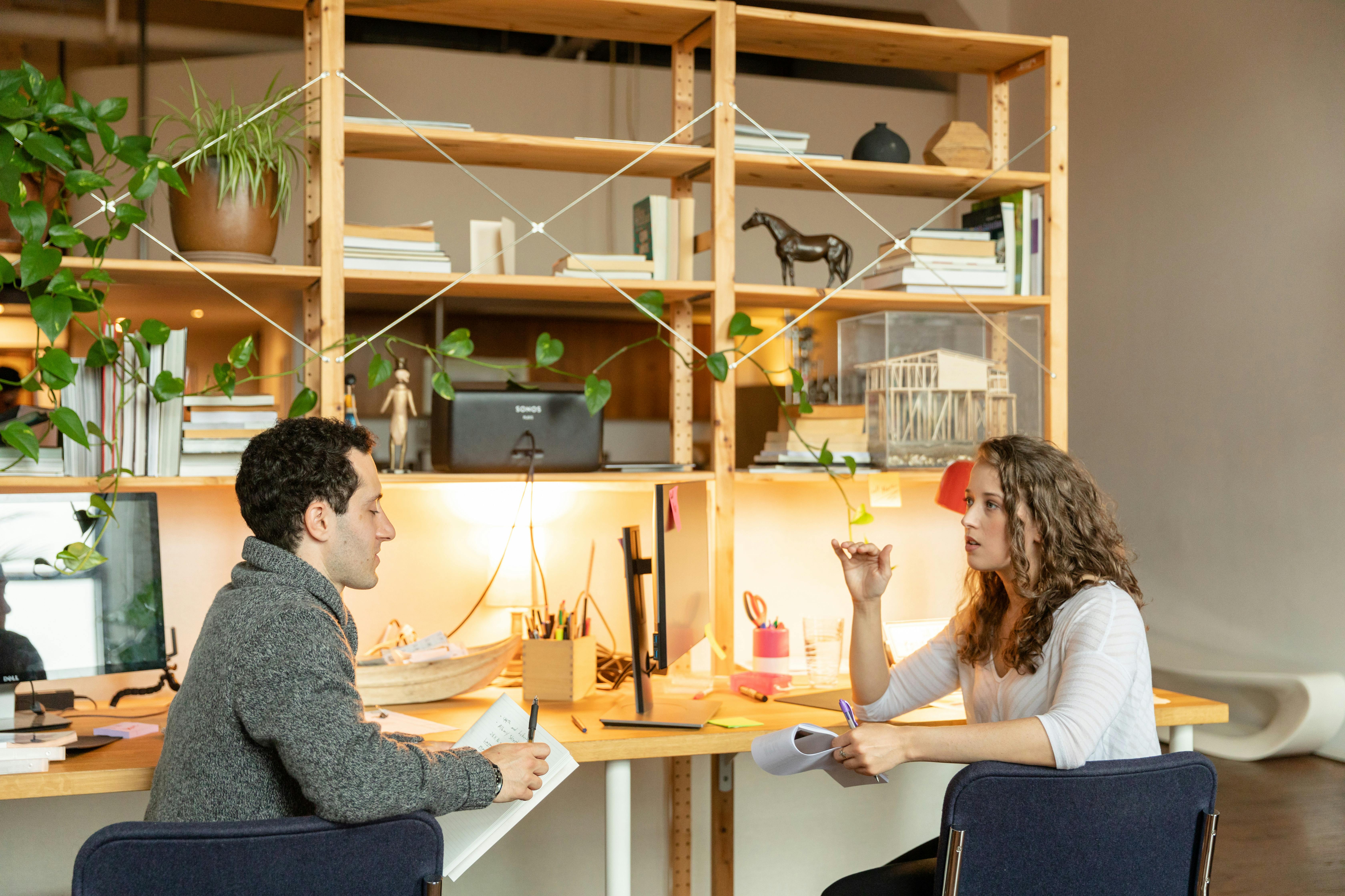 Two colleagues engaged in a discussion at a modern office desk with plants and books.