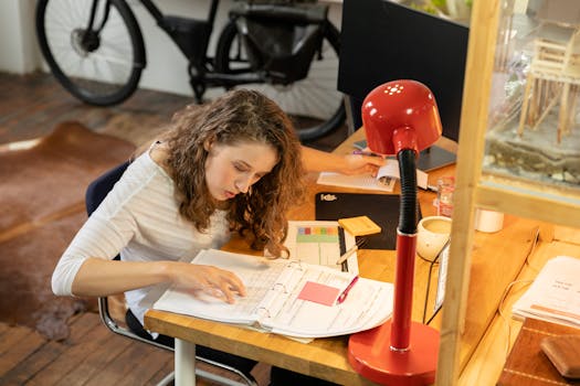 Young woman deeply focused on studying at a wooden desk with a red desk lamp.