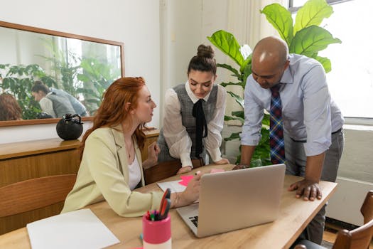 Three colleagues brainstorming around a laptop in a modern office setting.