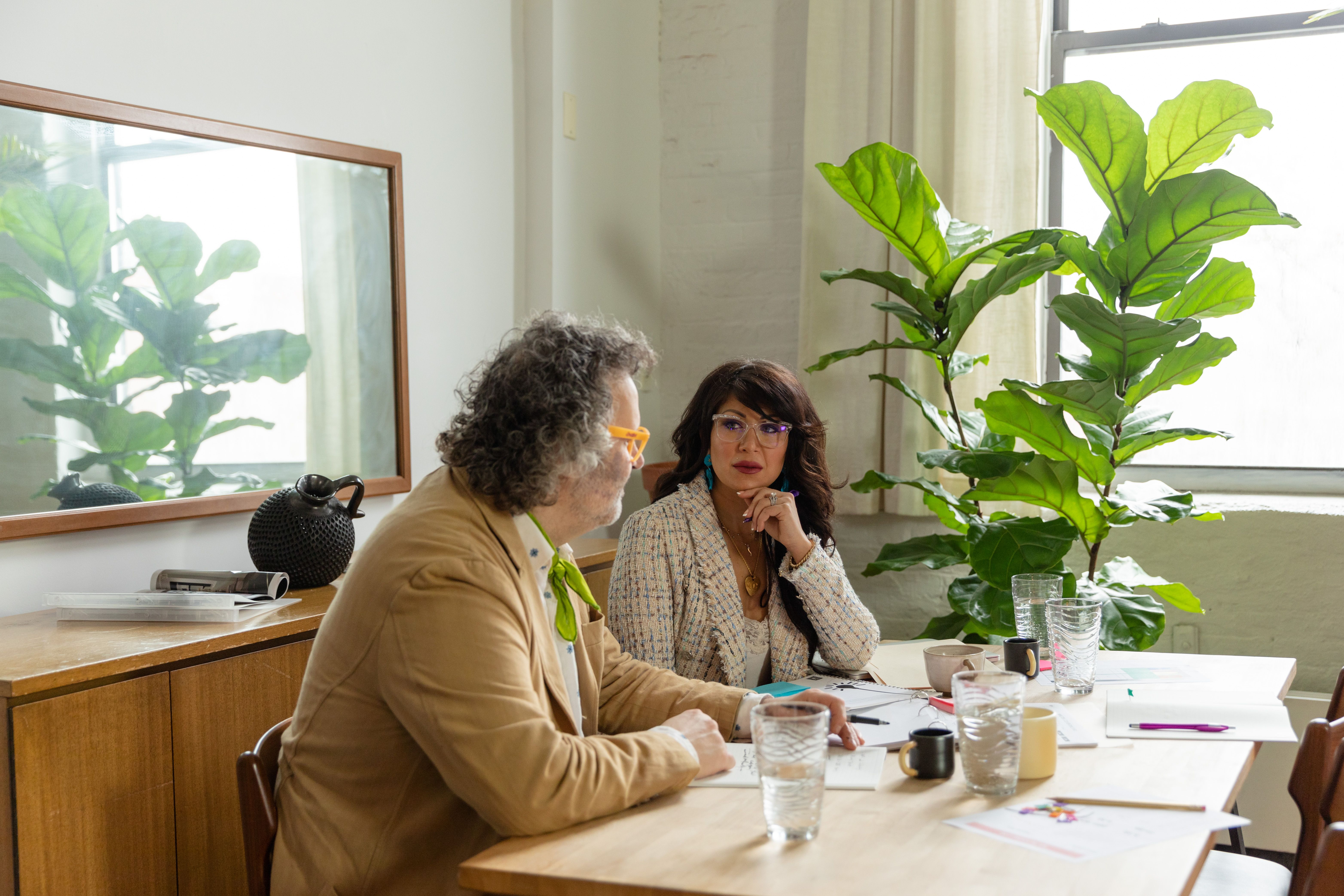 Two colleagues discussing work at a table in a plant-filled office. Bright and modern ambiance.