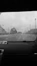 Black and white through glass POV shot of car driving in rain on asphalt road near buildings in city
