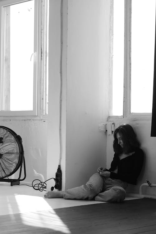 Black and white of upset brunette sitting alone on floor under window in room with shabby walls in daylight
