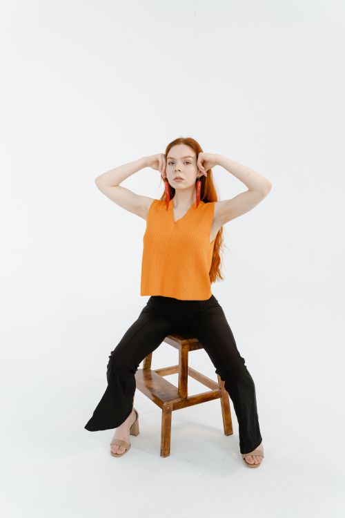 Free Confident woman in a bright orange top posing on a stool with hands near head. Stock Photo