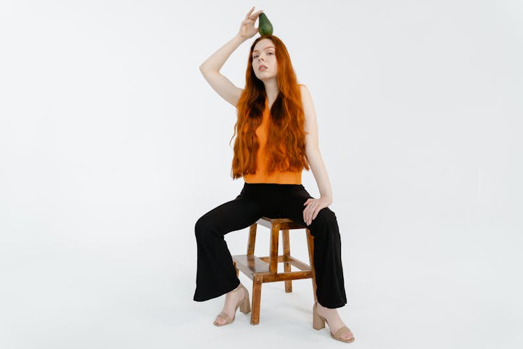 A Woman In Orange Top And Black Pants Holding An Avocado Fruit On Her Head