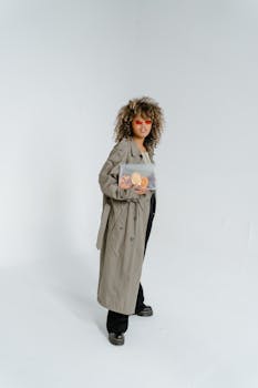 Fashionable woman with curly hair wearing trench coat and glasses, posing indoors.