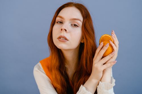 Free A woman with red hair holding a half orange against a blue background, showcasing beauty and freshness. Stock Photo