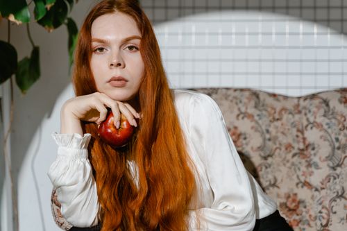 Free Elegant redhead woman holding an apple, posing in soft indoor lighting. Stock Photo