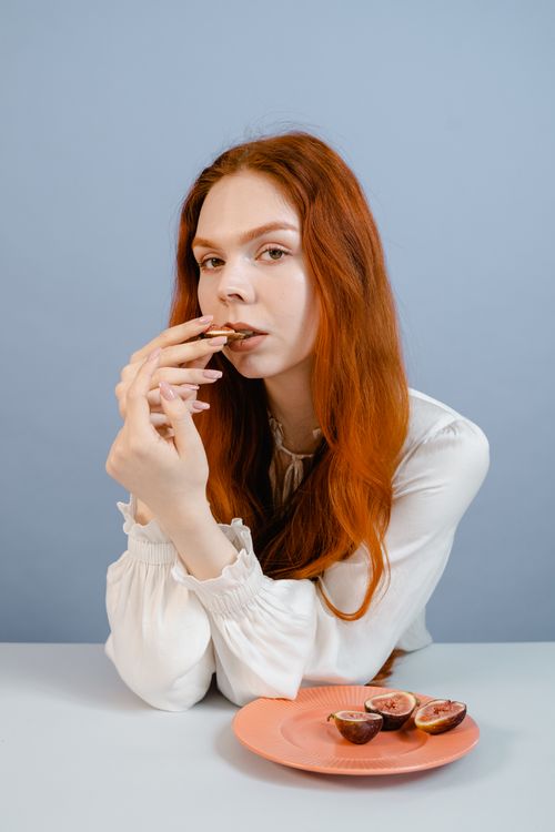 Free A redhead woman in white blouse savoring figs at a table with a pink plate against a blue background. Stock Photo