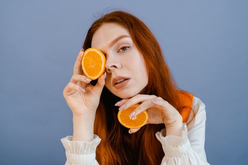 Free Vibrant portrait of a young woman with ginger hair holding orange slices against a blue background. Stock Photo