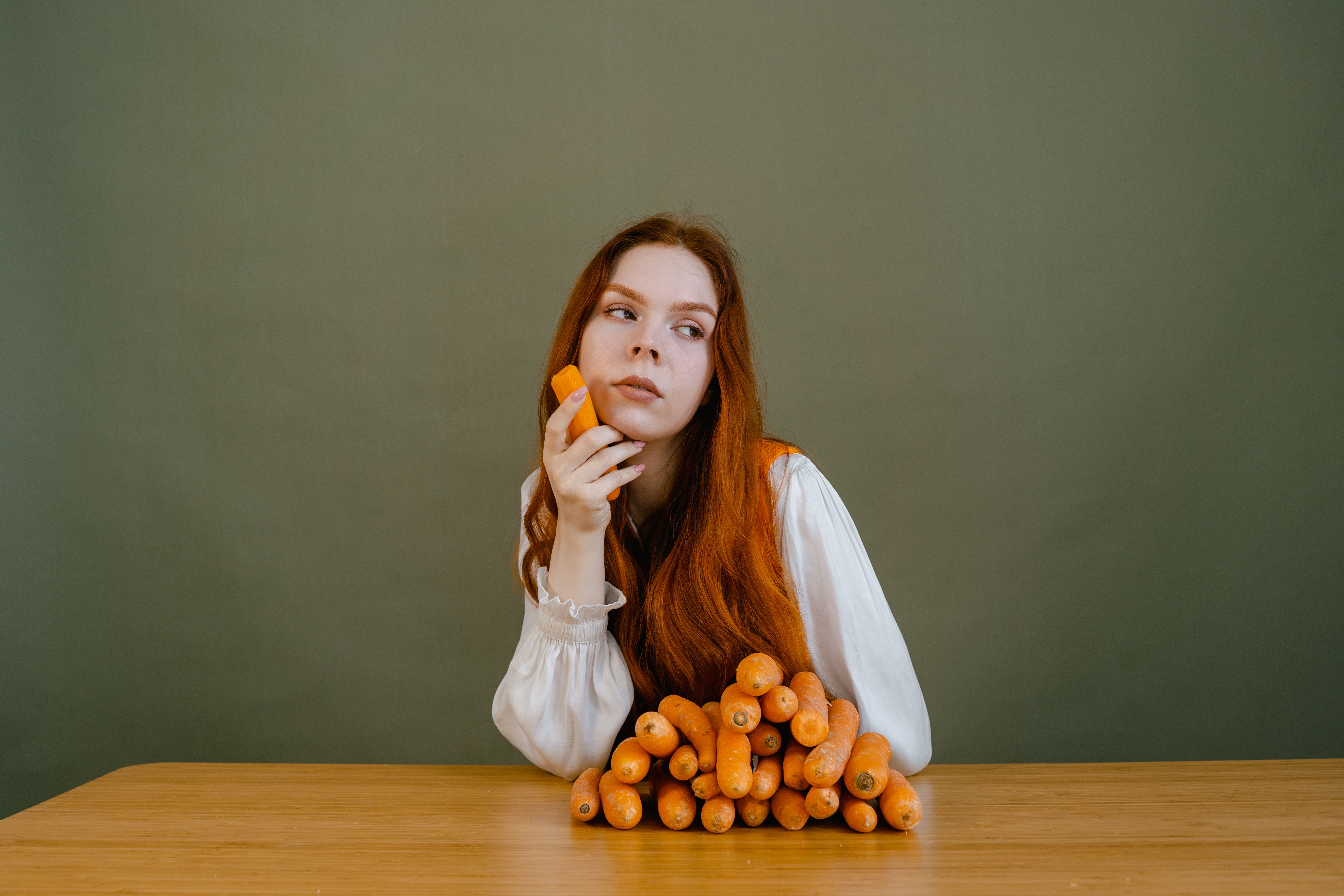 Red-haired woman in white sleeves holding fresh carrots on a wooden table against a green background.
