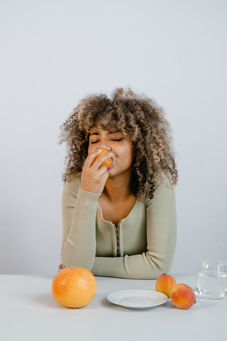 A Woman With Curly Hair Biting A Peach