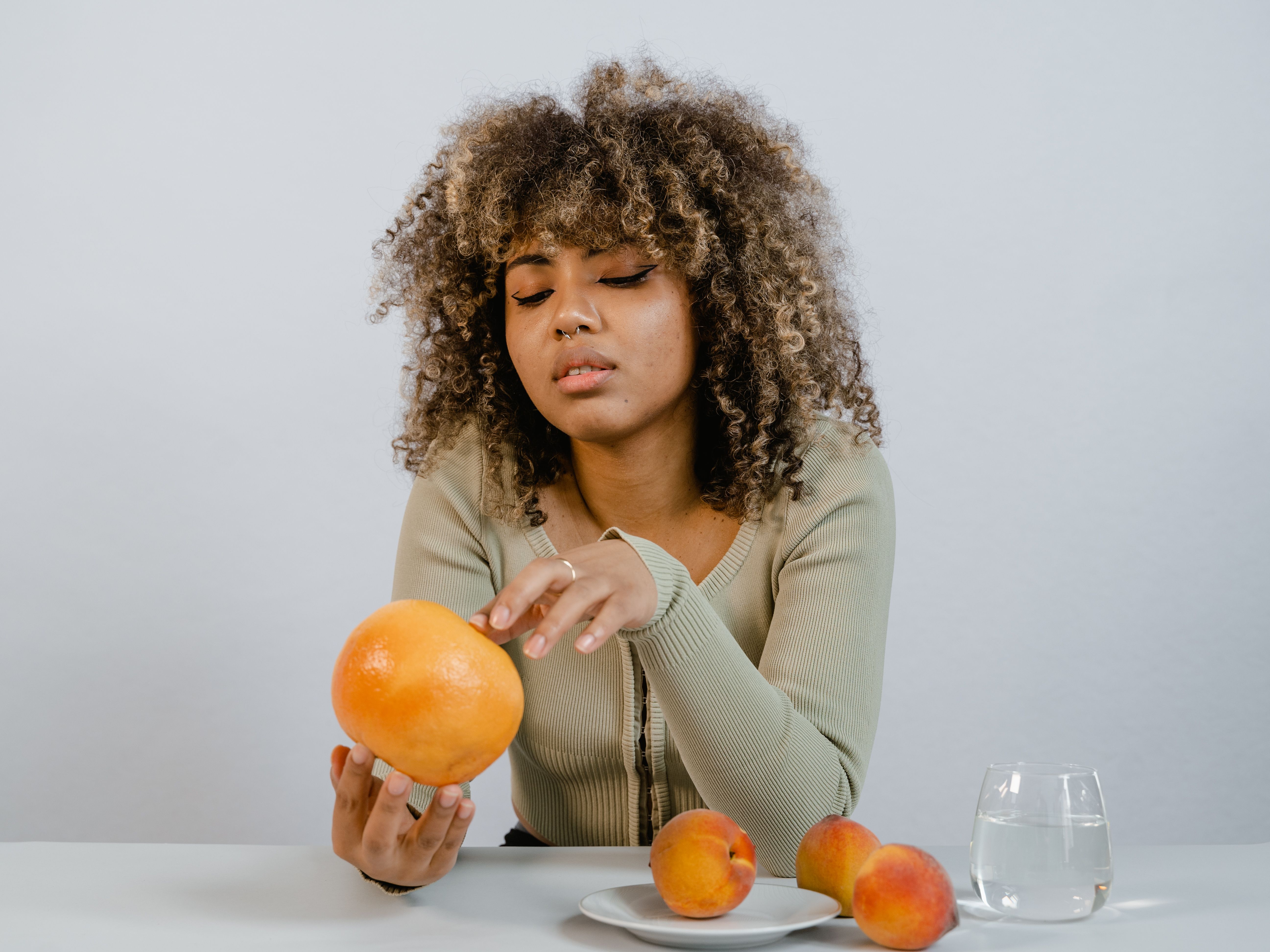 Woman with curly hair examines a citrus fruit on a table with peaches and water.