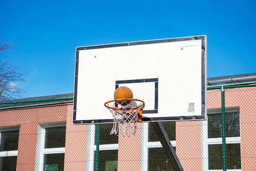 A basketball sits in a hoop against a clear blue sky on an outdoor court.