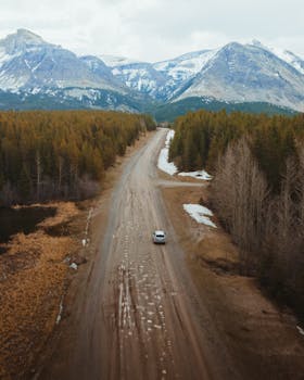 A car driving on a rural road amidst snow-capped mountains and forest, captured in an aerial view.