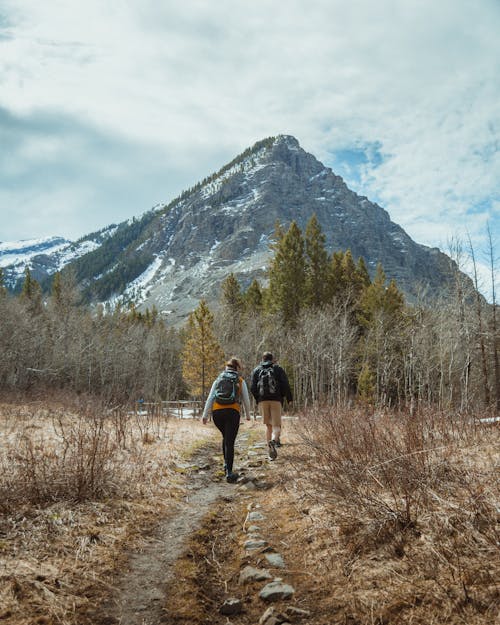 Two People with Backpacks Walking on Pathway Near Mountain