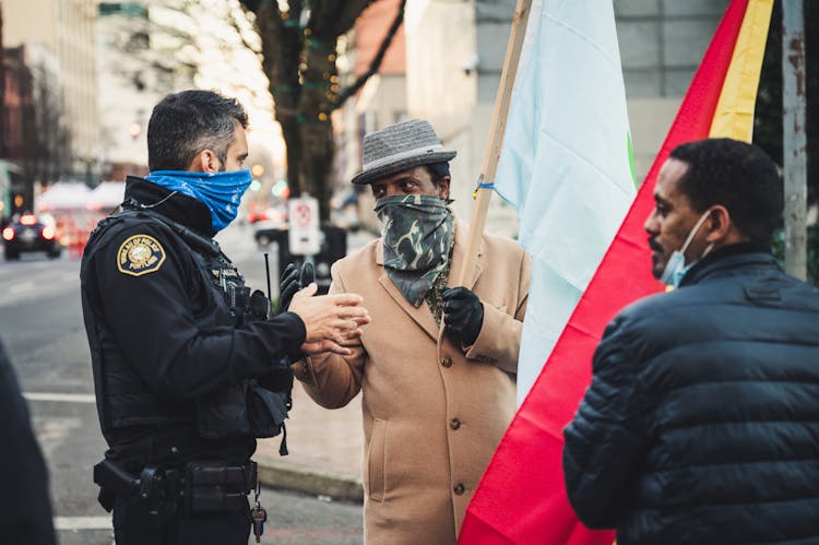 Policeman Standing And Talking With Black Men On City Street In Daytime