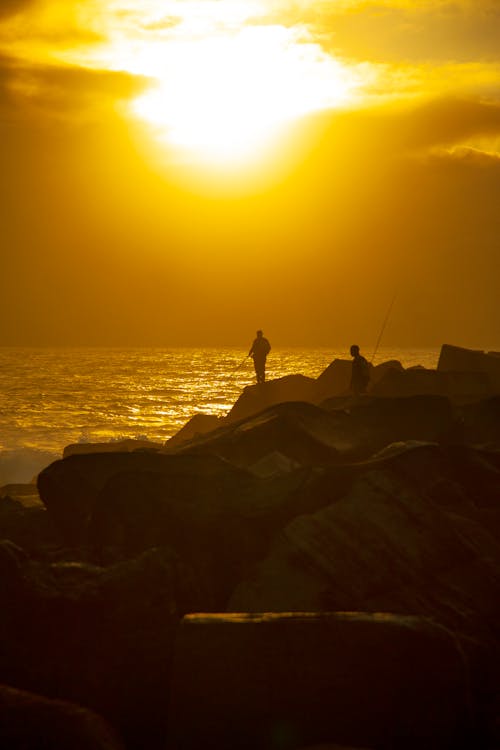 Silhouette of Persons With Fishing Rods on Rocks Near a Body of Water