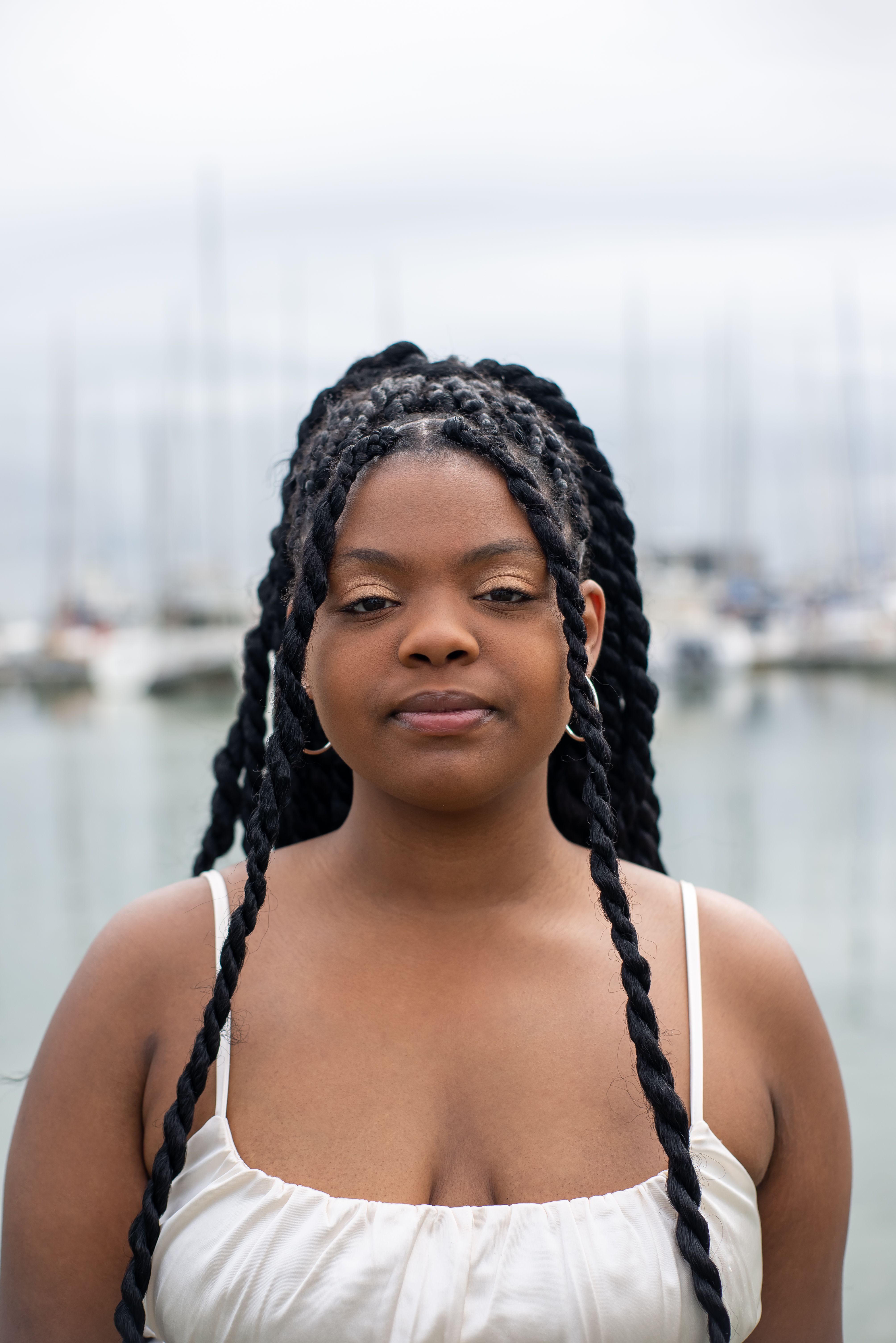A woman with braided hair posing by a marina, wearing a white top.
