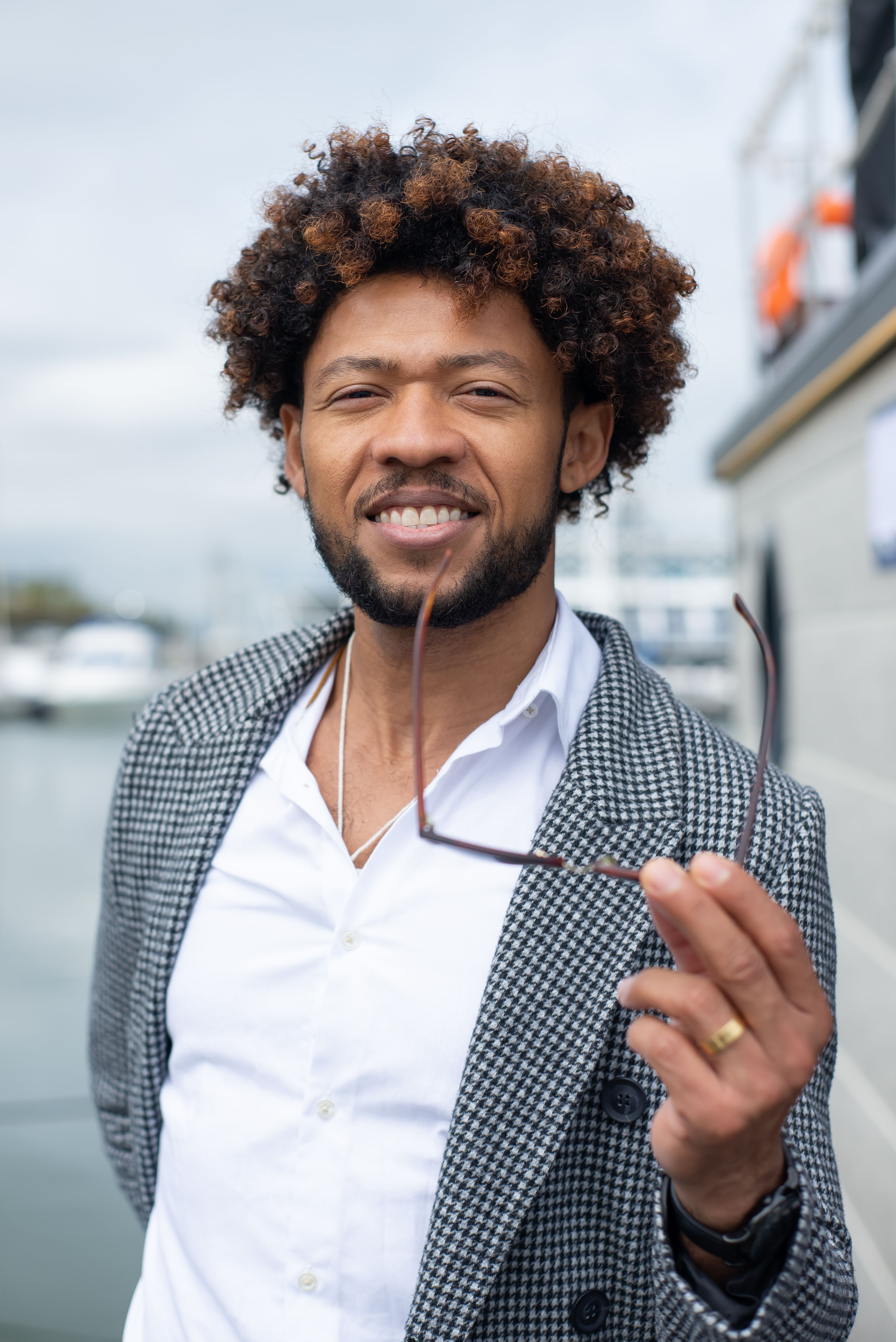 Confident man with afro hair holding eyeglasses, wearing a white shirt and blazer, smiling outdoors.