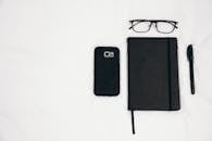 白色背景上摆放着黑色笔记本、智能手机、眼镜和笔，呈现出一个极简主义的平面布局