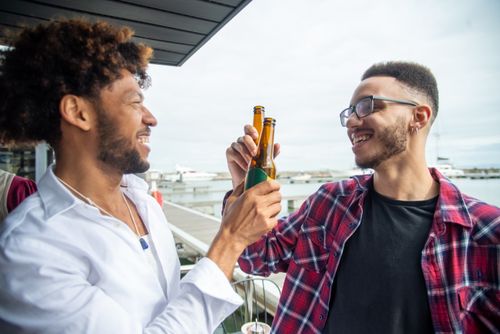 Free Two friends clinking beer bottles and smiling by a marina. Stock Photo