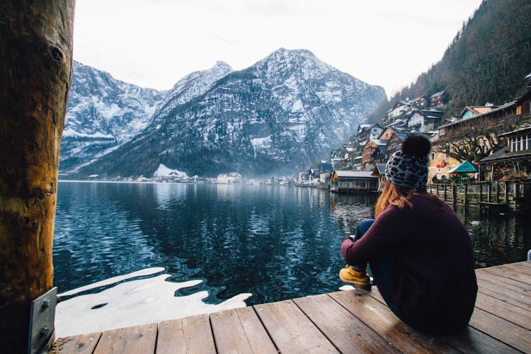 Woman In Purple Sweater Sitting On Wooden Floor With View Of Lake And Mountains