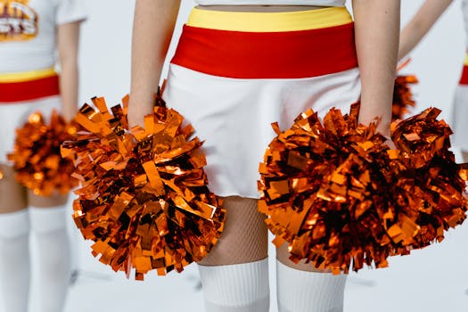 Close-up of a cheerleader holding bright orange pom poms, wearing a red and white uniform.