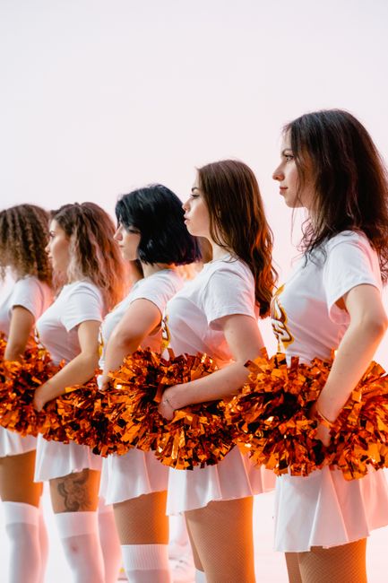 A line of cheerleaders in white uniforms with orange pompoms in a studio setting.
