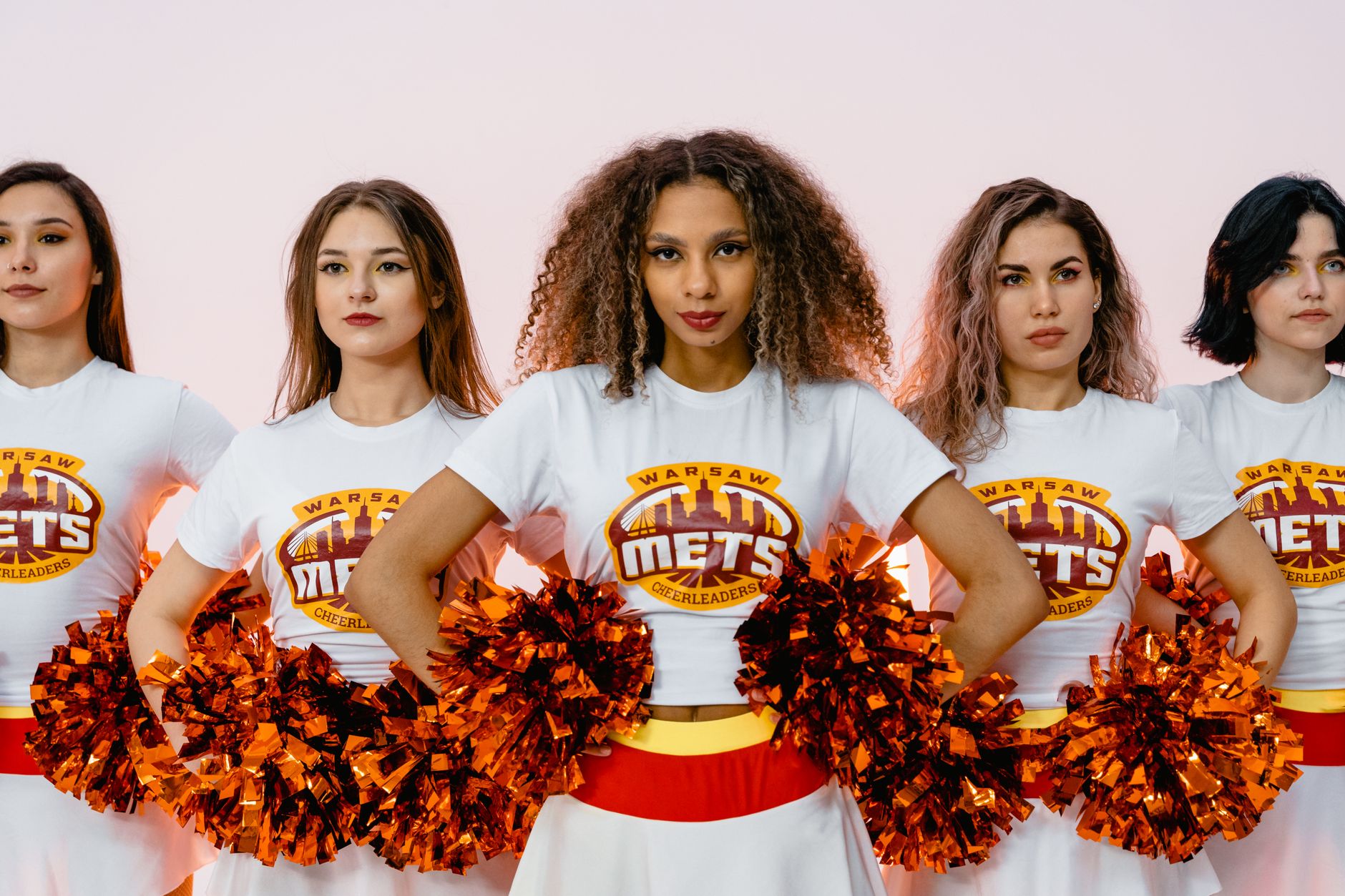 Group of diverse cheerleaders in white t-shirts with pom-poms on a pink background.