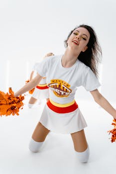 Cheerleader performing energetically indoors with bright orange pom poms and a white uniform.