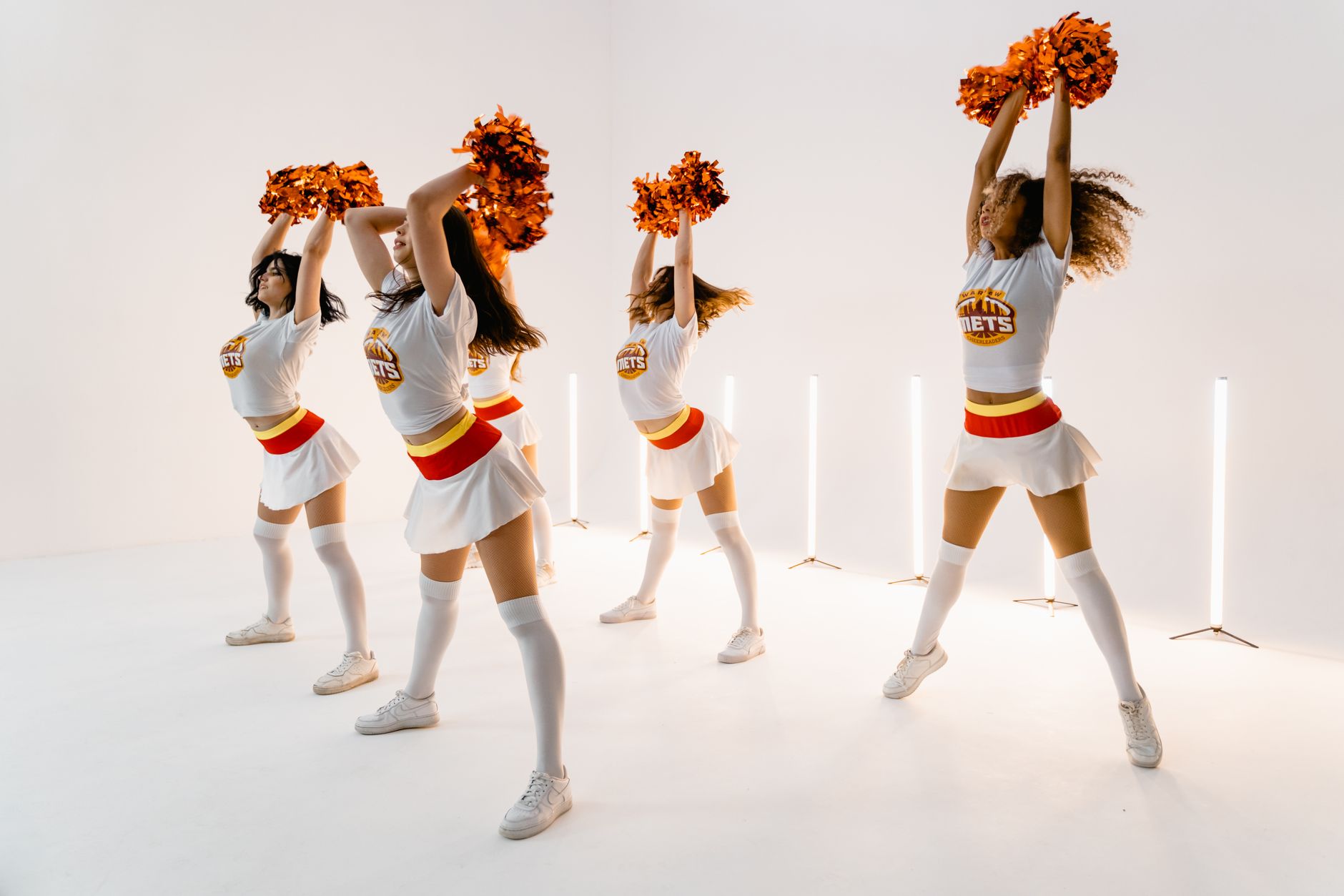 A vibrant group of cheerleaders performing choreography indoors with orange pompoms and uniforms.