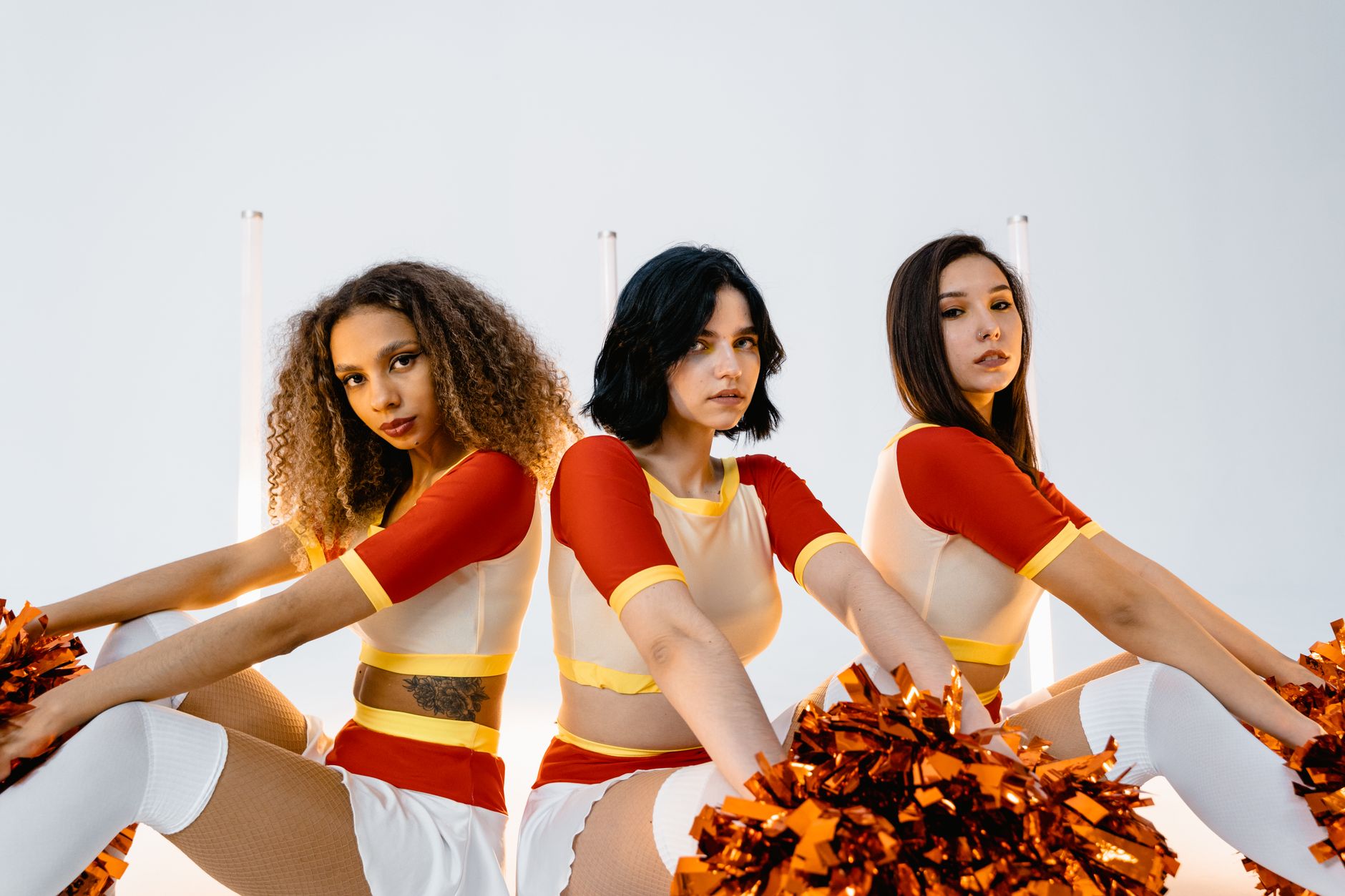 Energetic portrait of three diverse cheerleaders in vibrant uniforms holding pom poms indoors.