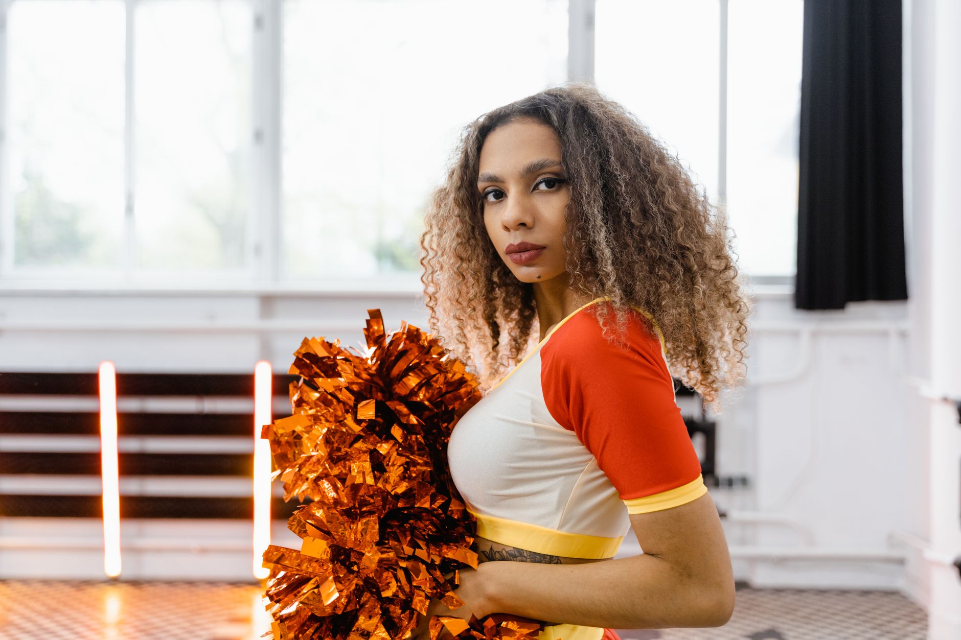 Young cheerleader holding vibrant orange pom poms in a gym setting, ready to perform.