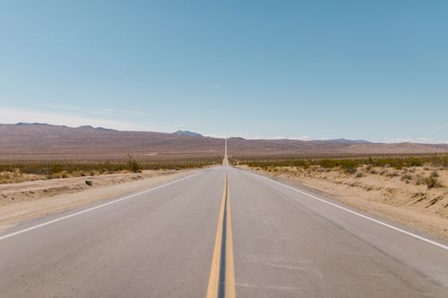 Free A long, straight road stretches through a vast desert landscape under a bright blue sky. Stock Photo