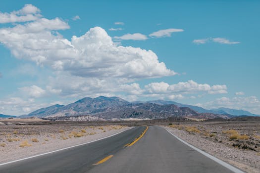 Empty highway stretches through arid landscape with mountains and blue sky.