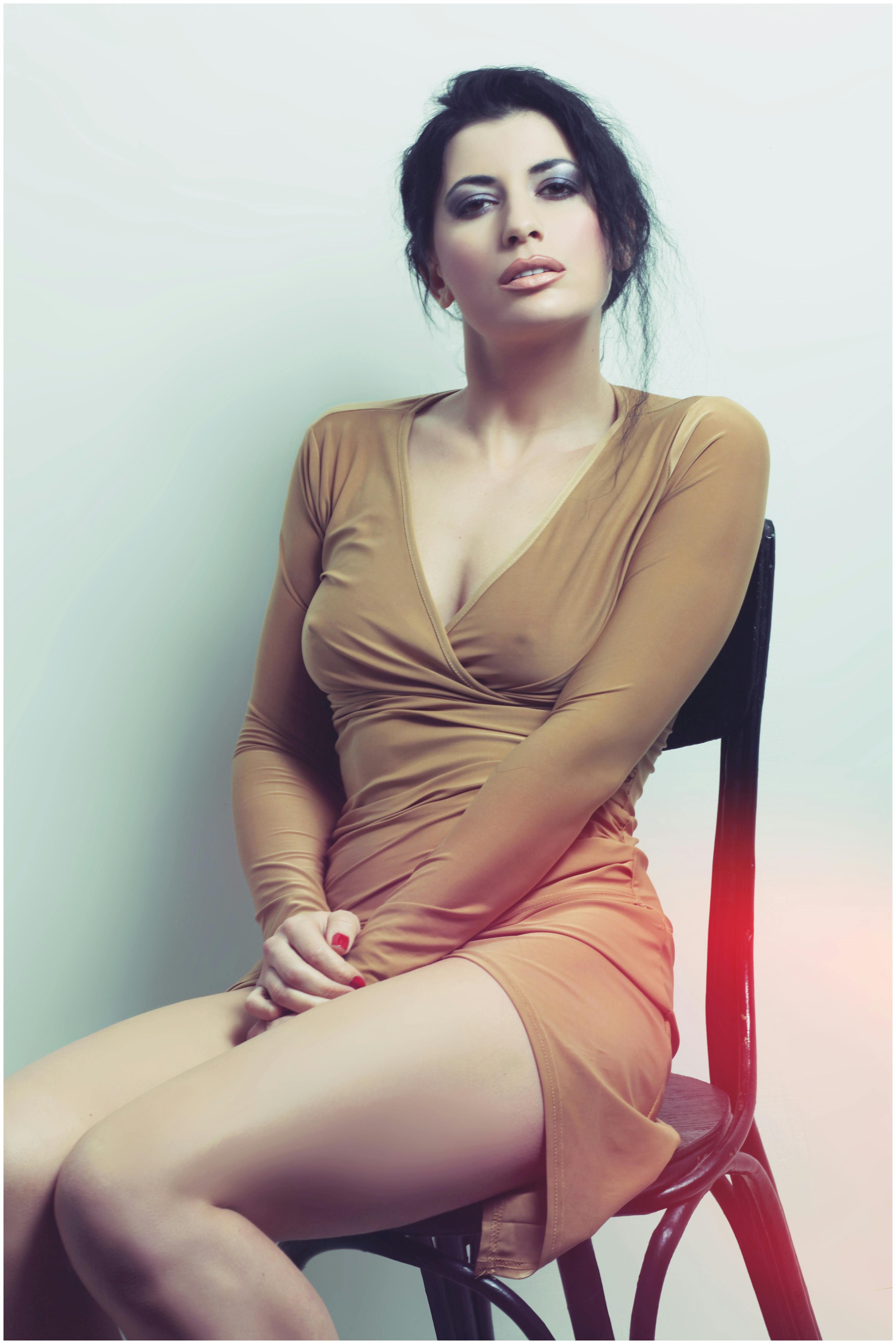 A fashionable woman poses confidently in a chic tan dress, seated on a chair.