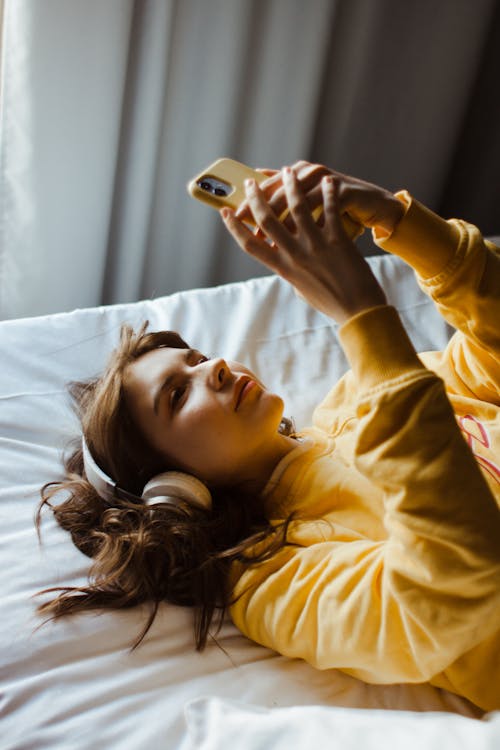 Free Young woman in a yellow sweater enjoying music on her phone while lying in bed. Stock Photo