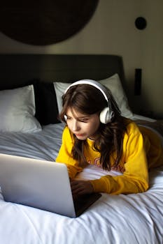 Woman wearing headphones, lying on bed, using laptop. Cozy indoor setting.