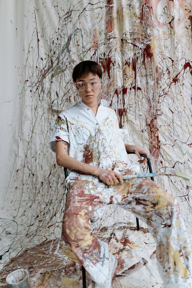 A Man With Paint On Clothes Sitting Near Messy Textile