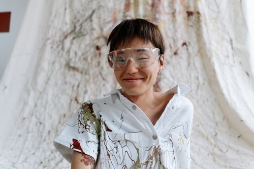 Free A cheerful Asian artist with short hair in safety glasses and a paint-splattered white shirt. Stock Photo