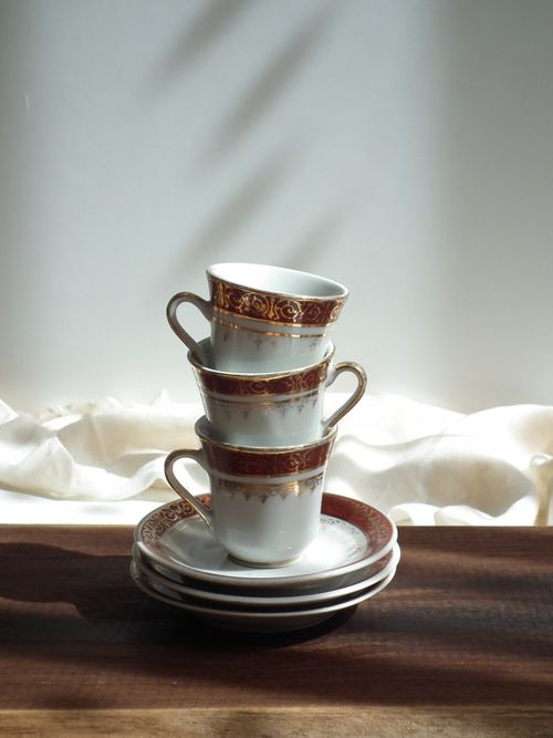 Free A stack of intricately designed porcelain tea cups with saucers on a wooden surface, lit by soft natural sunlight. Stock Photo