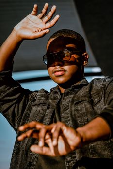 A confident young man in denim and sunglasses raises his hand against the bright sunlight, creating bold shadows.