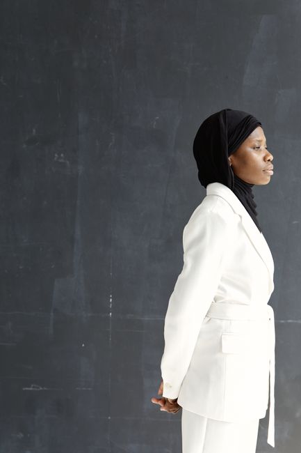 Stylish woman in a white suit and hijab posing side view against a dark background.