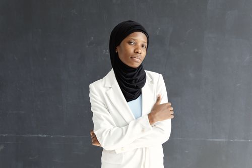 Free A confident Muslim woman poses in a white blazer and black hijab against a chalkboard background. Stock Photo