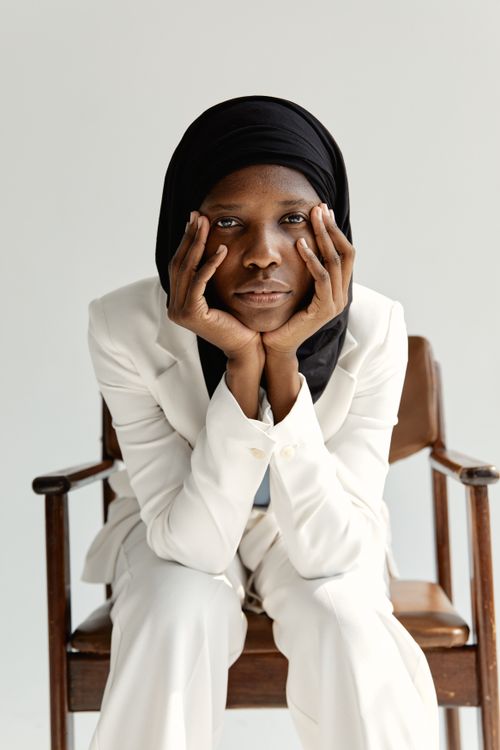 Free Elegant portrait of a woman in a white suit and hijab sitting thoughtfully on a chair. Stock Photo