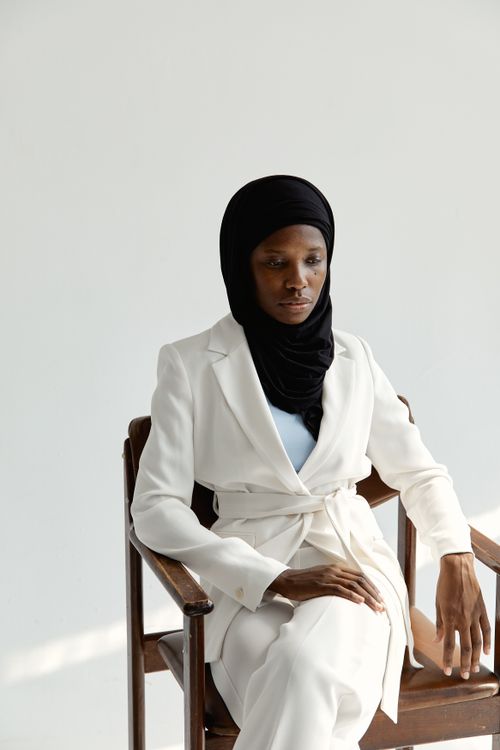 Free Portrait of a woman wearing a white suit and hijab sitting on a wooden chair against a white background. Stock Photo