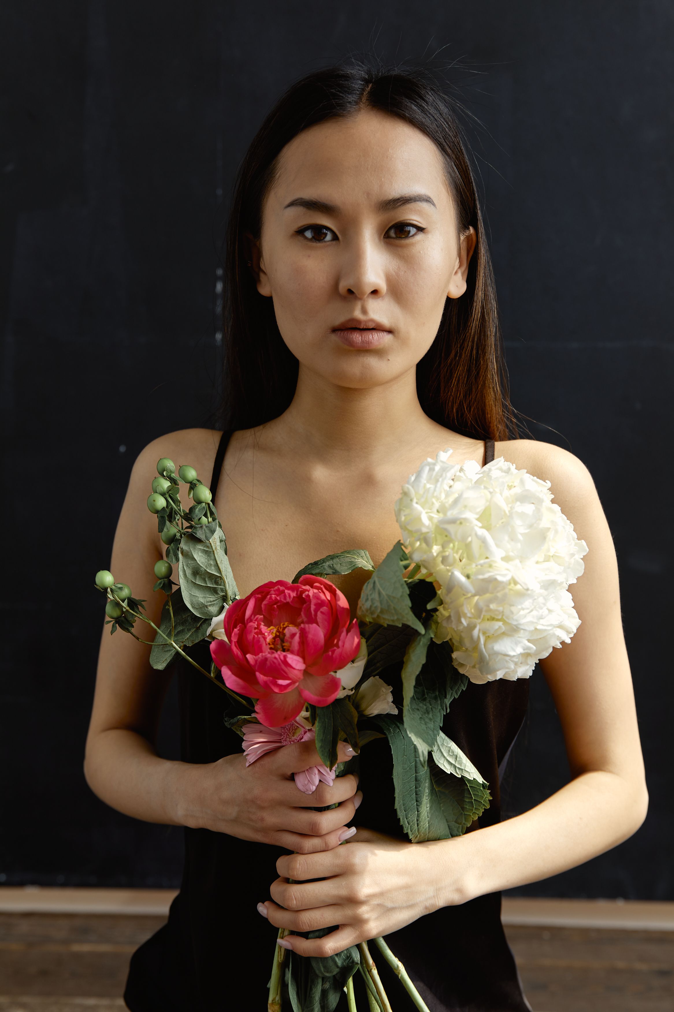 Asian woman in black dress holding peony and hydrangea against dark background